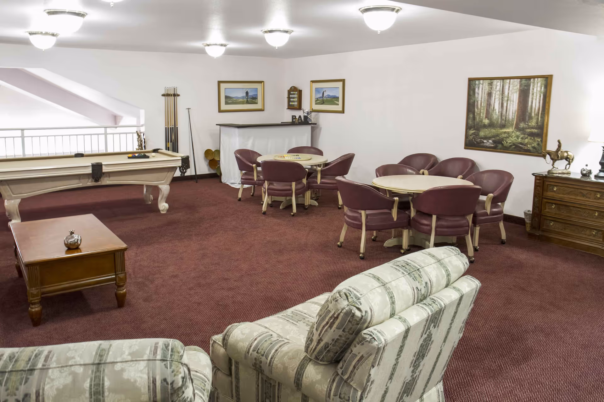 A communal lounge with a pool table, round card tables and upholstered chairs on burgundy carpet.