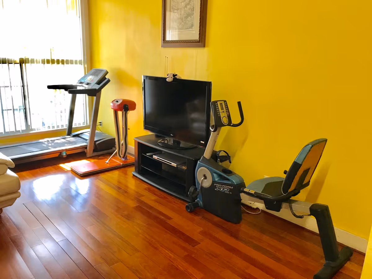 A room with bright yellow walls and wooden flooring featuring exercise equipment including a treadmill and a recumbent exercise bike, a black TV on a stand, and a red vibration plate machine near a large window with vertical blinds.