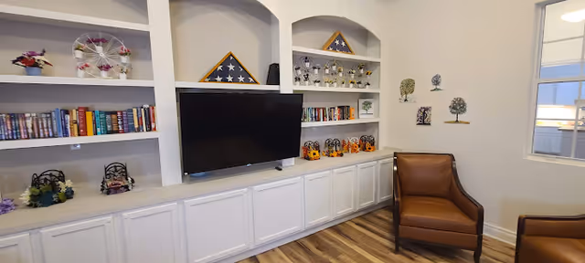 A cozy living room area with built-in white shelves filled with books, decorative items, and two folded American flags in triangular display cases. A flat-screen TV is mounted on the shelves. There are two brown leather armchairs on a wood floor, and a window on the right side lets in natural light. The walls are decorated with small tree-themed wall art.