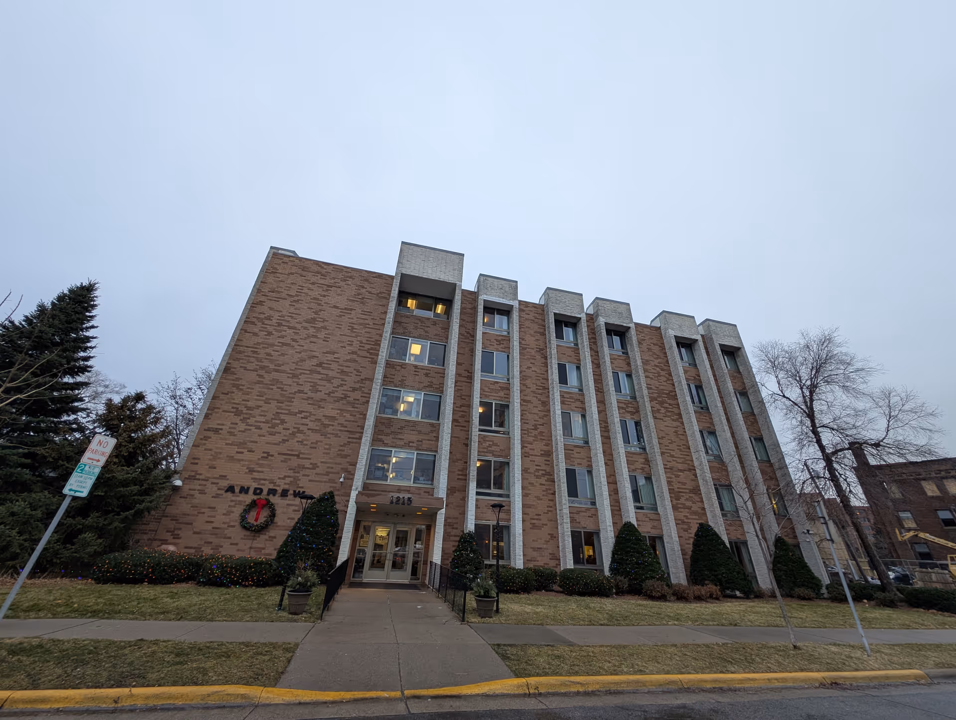 Exterior view of a multi-story brick building named Andrew Residence with several windows, a main entrance with glass doors, and some landscaping including bushes and trees. The sky is overcast.