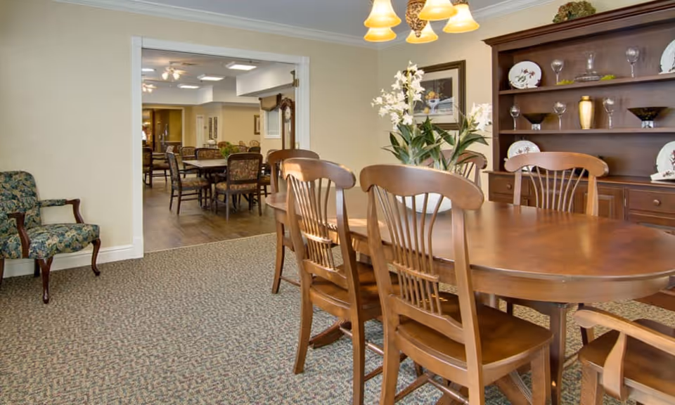 Wood-furnished dining room with a long table and chairs, a china hutch, floral centerpiece, and a view into an adjoining dining area.