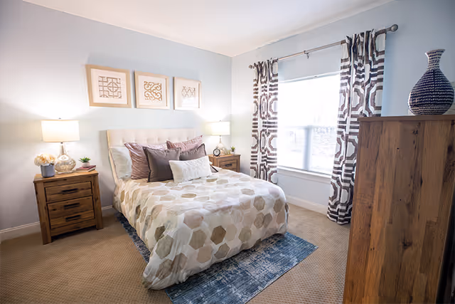 A cozy bedroom with a bed centered against a light blue wall, featuring a beige upholstered headboard and a bedspread with hexagonal patterns in neutral tones. There are multiple pillows on the bed. On each side of the bed, there are wooden nightstands with lamps and decorative items. A large window with patterned curtains allows natural light to fill the room. A wooden dresser with a decorative vase is positioned on the right side of the room, and a blue rug is partially under the bed on a beige carpeted floor.