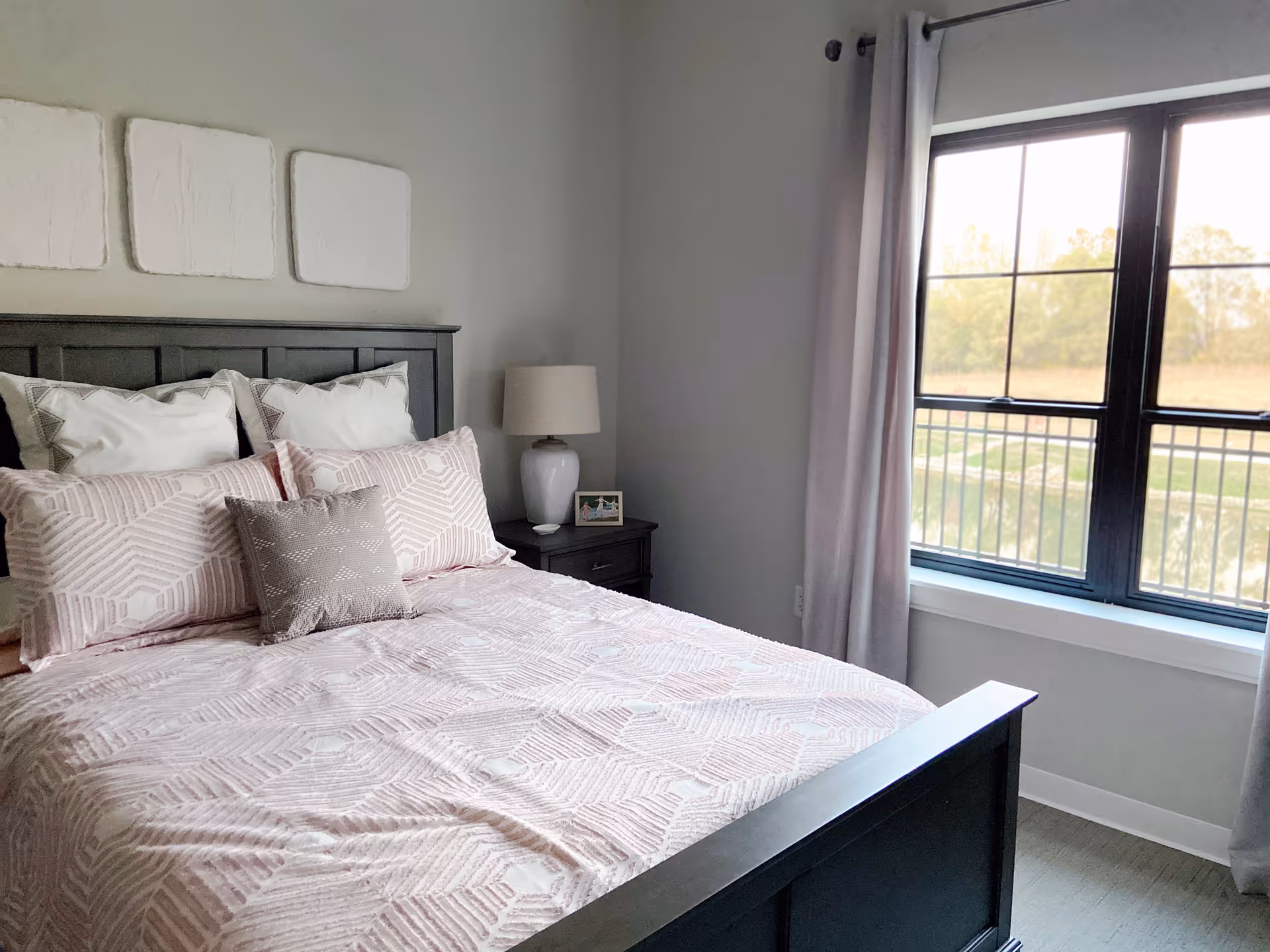 Sunlit bedroom with a dark wood bed dressed in pink patterned bedding, a nightstand with a lamp, and a large window overlooking the outdoors.