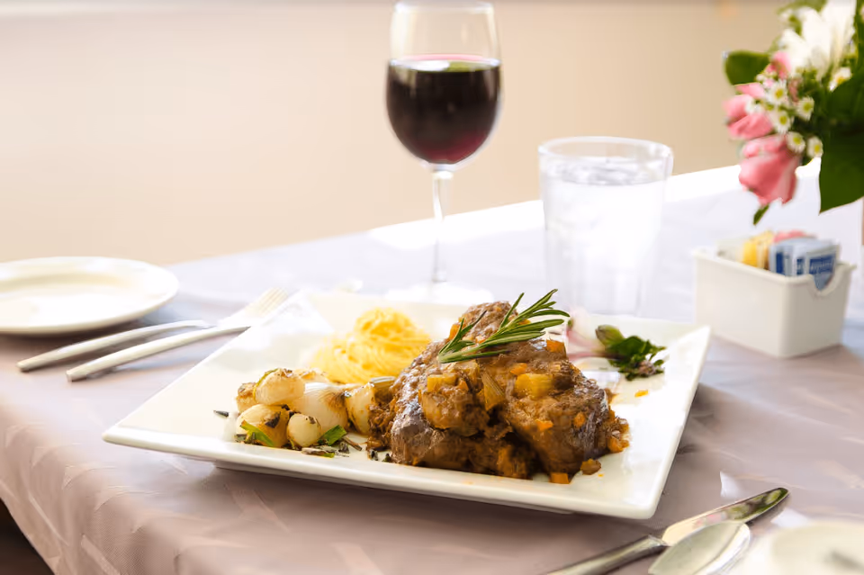A plated meal consisting of cooked meat garnished with a sprig of rosemary, accompanied by roasted onions and a serving of mashed potatoes. The plate is on a table set with a glass of red wine, a glass of water with ice, silverware, and a small container holding sugar packets. A small bouquet of pink and white flowers is also visible on the table.