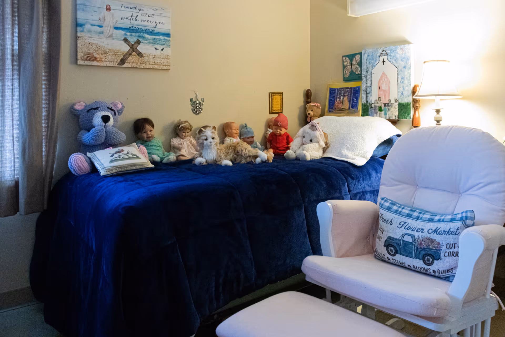 Cozy bedroom with a navy-covered bed lined with dolls and stuffed animals, a pink armchair with a decorative pillow, and a lamp.