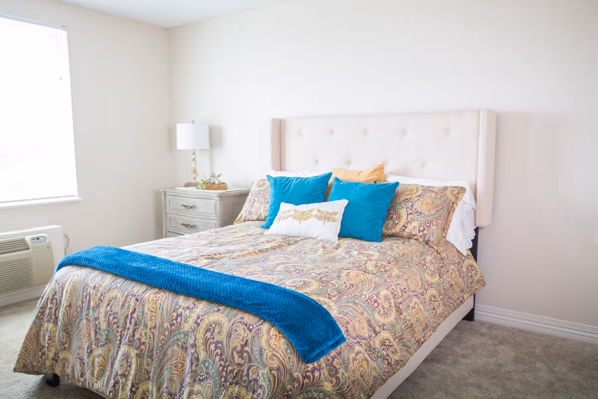A bright bedroom with a large bed featuring a beige tufted headboard, paisley-patterned bedding in shades of gold and purple, and several decorative pillows in blue, white, and gold. There is a small nightstand with a lamp and decorative items next to the bed, and a window with blinds letting in natural light.