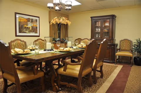 A formal dining room with a large wooden dining table set for a meal, surrounded by eight upholstered chairs. The table has place settings with plates, glasses, napkins, and decorative candles. A wooden china cabinet is against the wall, and a framed painting hangs above a curtain in the background. The room has warm lighting and a patterned carpet.