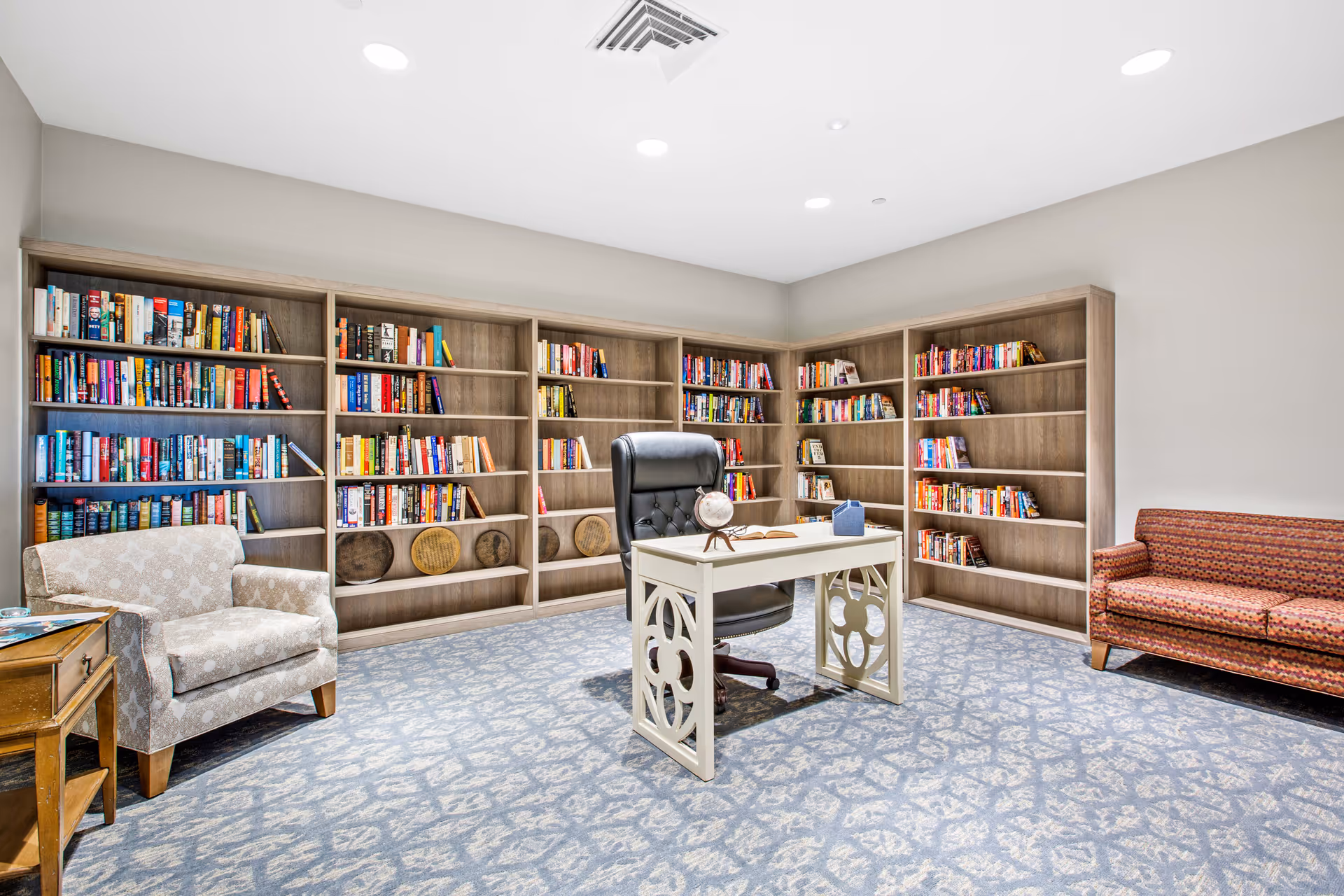 A bright and spacious library room with light gray walls and patterned carpet. The room features multiple wooden bookshelves filled with colorful books along the back and right walls. In the center, there is a white decorative desk with a black leather office chair. On the left side, there is a beige patterned armchair next to a small wooden side table, and on the right side, a red patterned sofa.