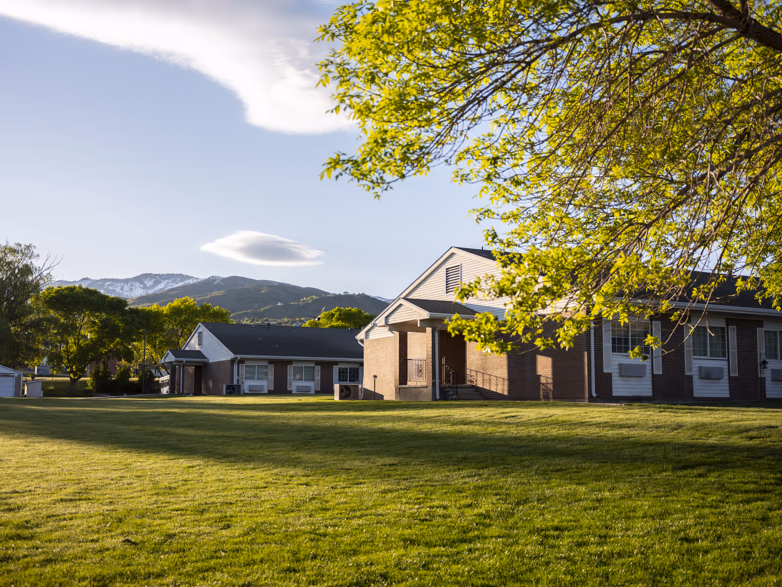 Exterior view of a single-story brick building with white trim, surrounded by a large green lawn and trees, with mountains visible in the background under a clear sky.