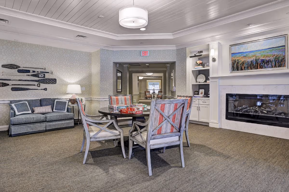 Cozy senior living common room with a round table and chairs, a sofa, fireplace, and decorative wall art.