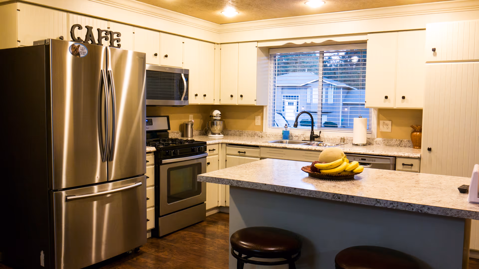 A modern kitchen with stainless steel refrigerator, oven, and microwave. White cabinets with black knobs surround the kitchen. A window above the sink shows a view of neighboring houses. A kitchen island with a granite countertop has a bowl of bananas and a melon on it, with two dark round stools underneath.