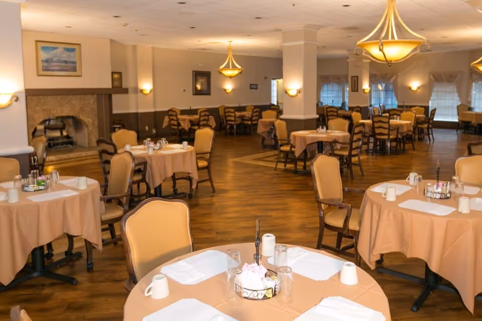 Large dining room with round tables set with place settings and beige chairs in a senior living facility.
