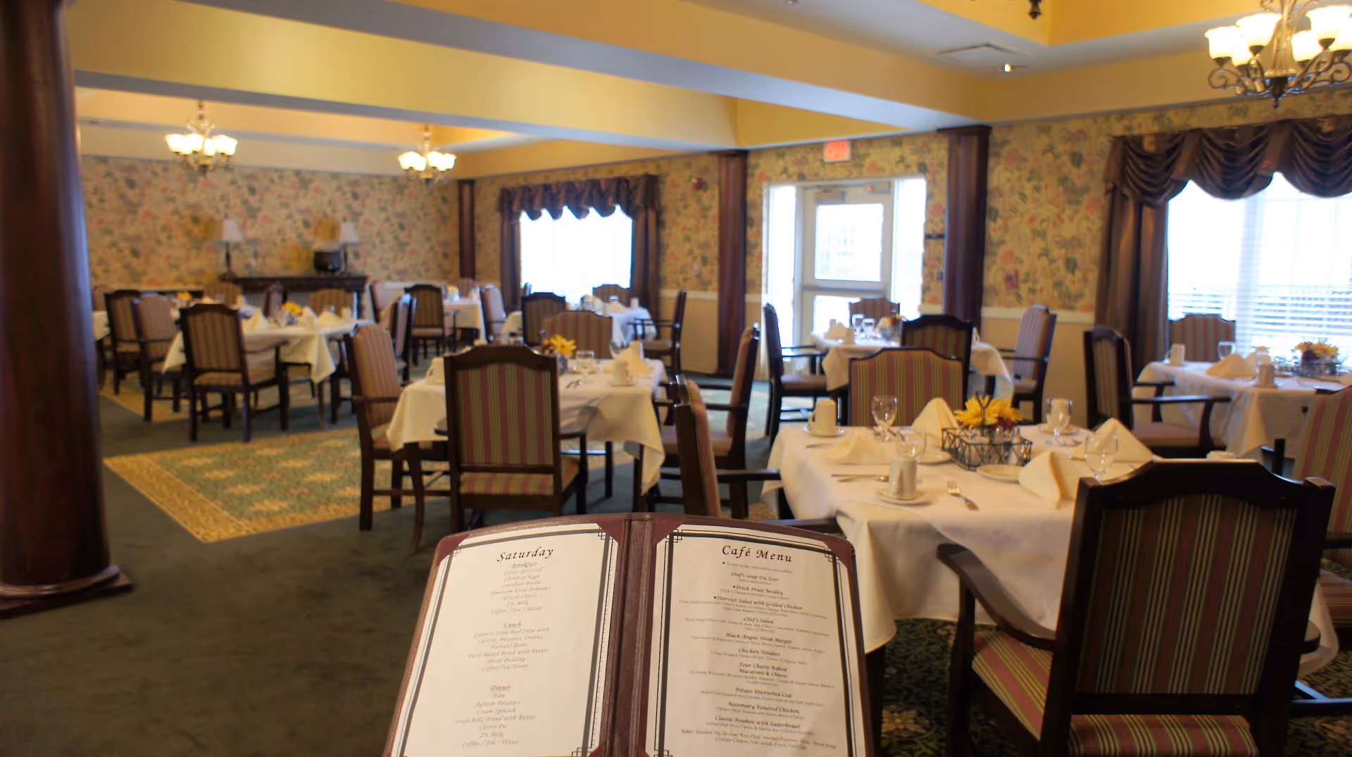 Interior view of a dining room in a senior living facility with multiple tables covered in white tablecloths, set with napkins, glasses, and silverware. The room has floral wallpaper, carpeted floors, and large windows with drapes allowing natural light. A menu is visible in the foreground.