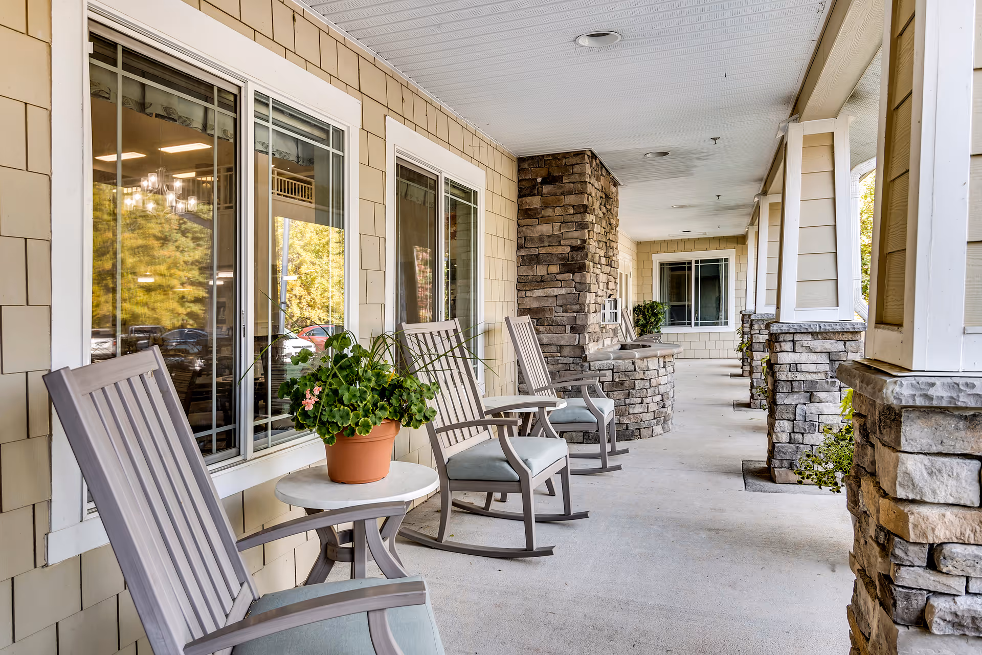 A covered outdoor porch area with several wooden rocking chairs and small round tables. One table has a potted plant with green leaves and pink flowers. The porch features stone pillars and beige siding with large windows reflecting trees and parked cars.