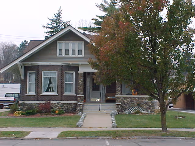 Front exterior view of a single-story brick house with a stone foundation, a small porch with steps, and a tree partially obscuring the right side of the house. The house has multiple windows with white trim and a gabled roof.