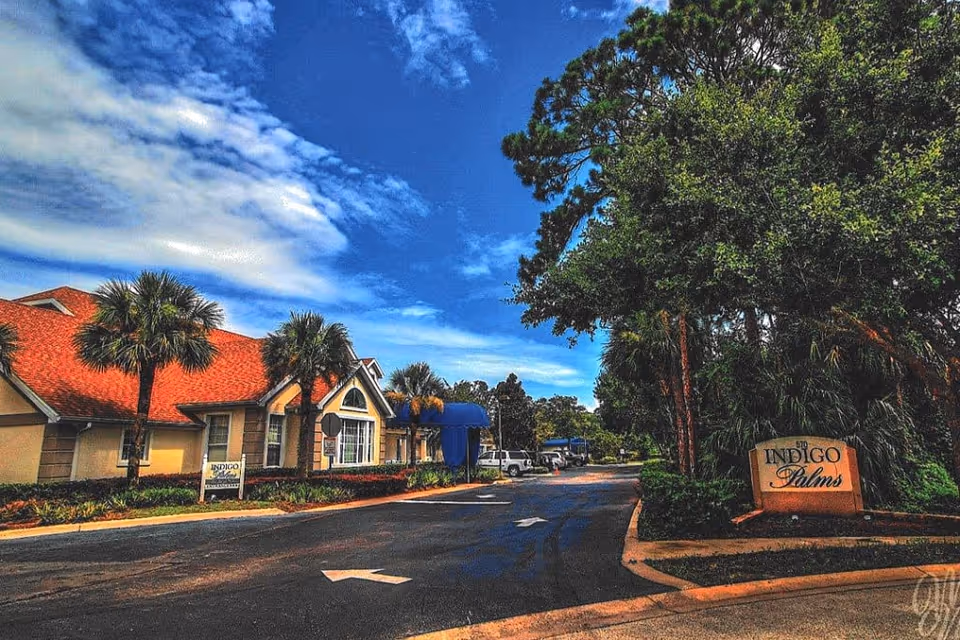 Entrance driveway and front of the Indigo Palms community with its sign, palm trees, and a low building under a blue sky.