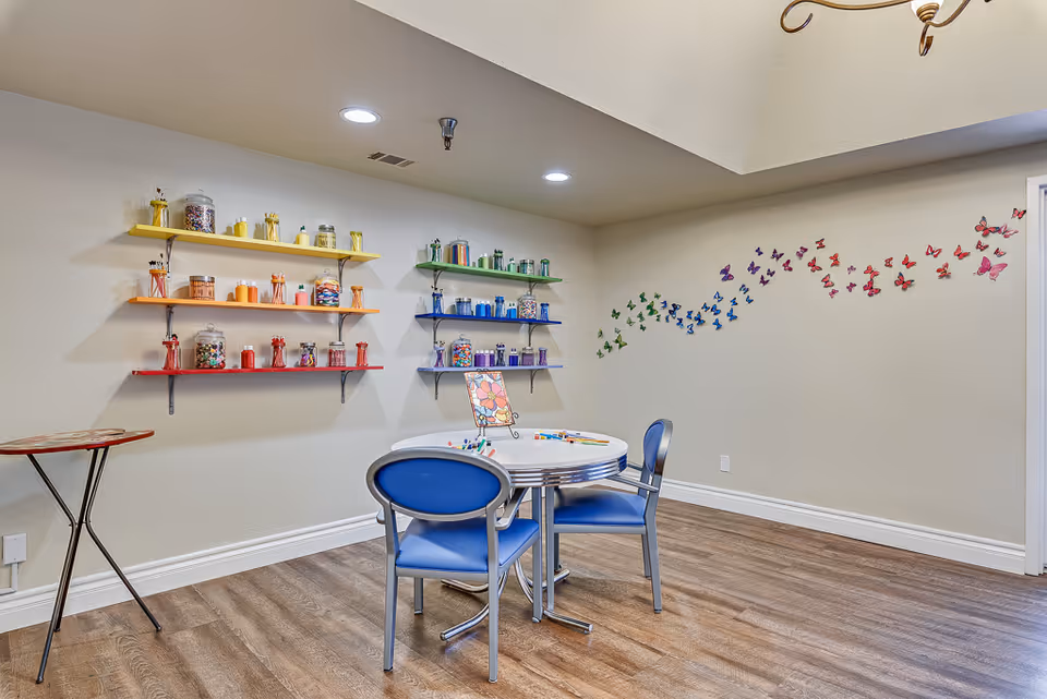 A bright and colorful activity room with a round table and two blue chairs. The walls feature shelves painted in rainbow colors holding jars filled with various craft supplies. A wall decoration of butterflies in a rainbow pattern is displayed on the adjacent wall. The room has wood flooring and recessed ceiling lights.