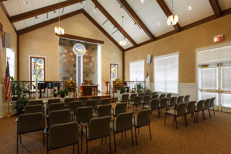 Interior view of a chapel or worship room with rows of chairs facing a raised platform with a pulpit, floral arrangements, and stained glass windows. The room has a high vaulted ceiling with wooden beams and hanging light fixtures. There are large windows with blinds and an exit door on the right side.
