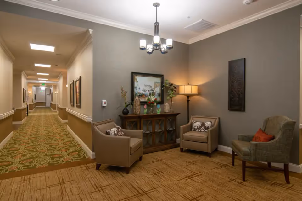 A cozy seating area in a senior living facility hallway with three armchairs arranged around a wooden cabinet adorned with decorative items and a framed painting above it. The walls are painted in neutral tones, and a floor lamp and ceiling light fixture provide warm lighting. The hallway extends to the left with patterned carpet and framed artwork on the walls.