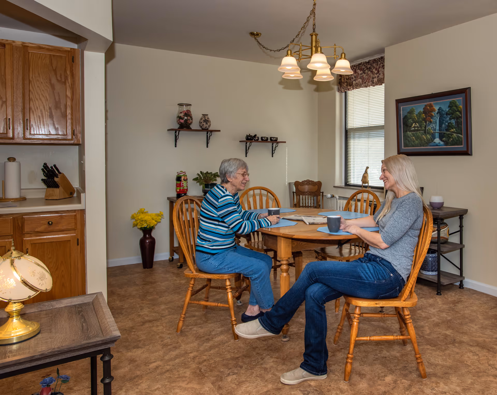 Two women seated at a wooden dining table in a cozy dining area, talking and holding mugs.