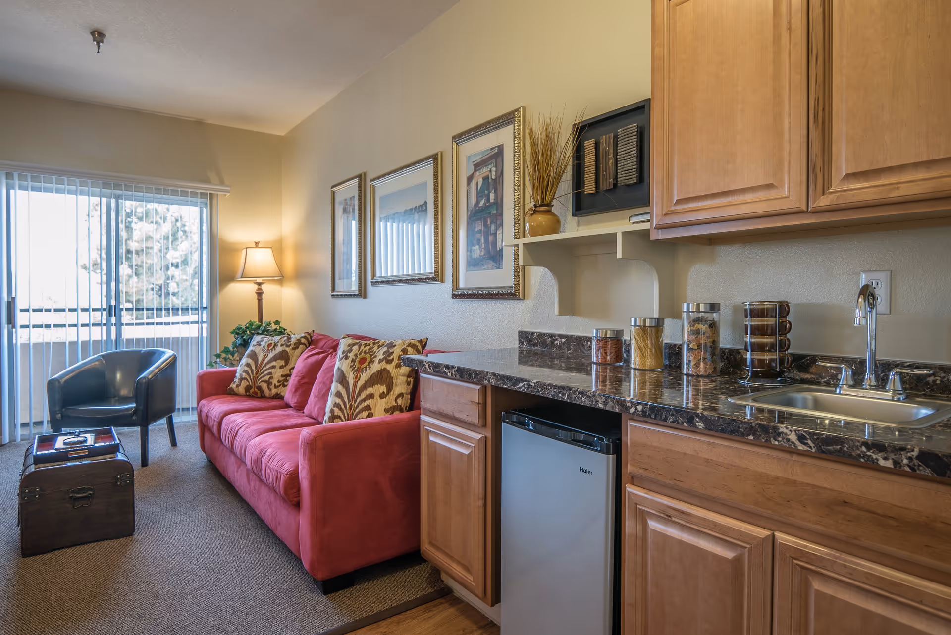 A cozy living area featuring a red sofa with patterned cushions, a black armchair, and a small wooden chest used as a coffee table. The room has a sliding glass door with vertical blinds letting in natural light. On the right side, there is a kitchenette with wooden cabinets, a small refrigerator, a sink, and several jars with food items on the marble countertop. The walls are decorated with framed artwork and a decorative vase with dried plants.