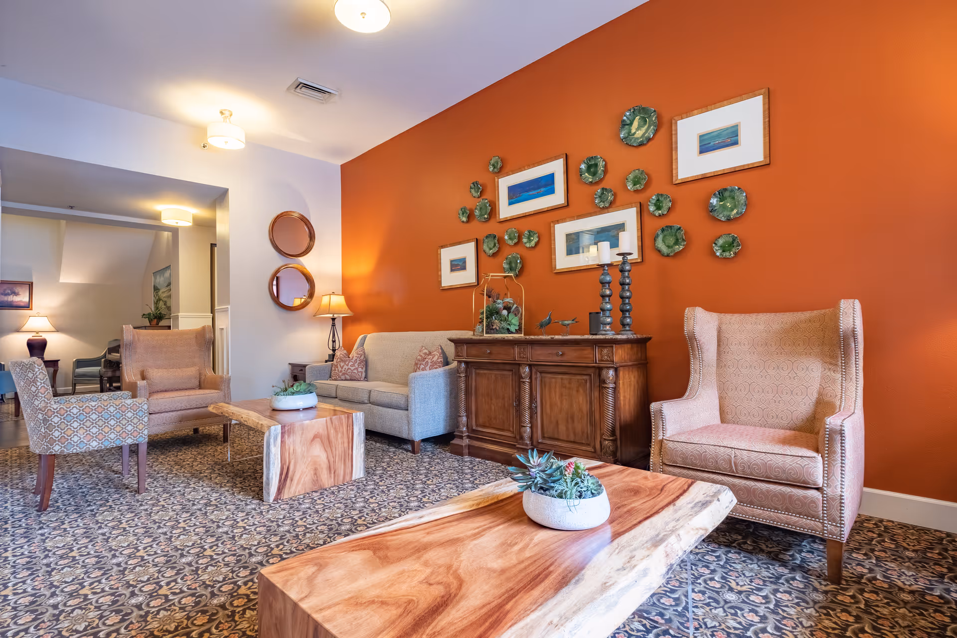 A cozy living room area with a patterned carpet, a wooden coffee table with a small plant, a beige sofa, two upholstered armchairs, and a wooden sideboard against an orange accent wall decorated with framed pictures and green decorative plates.