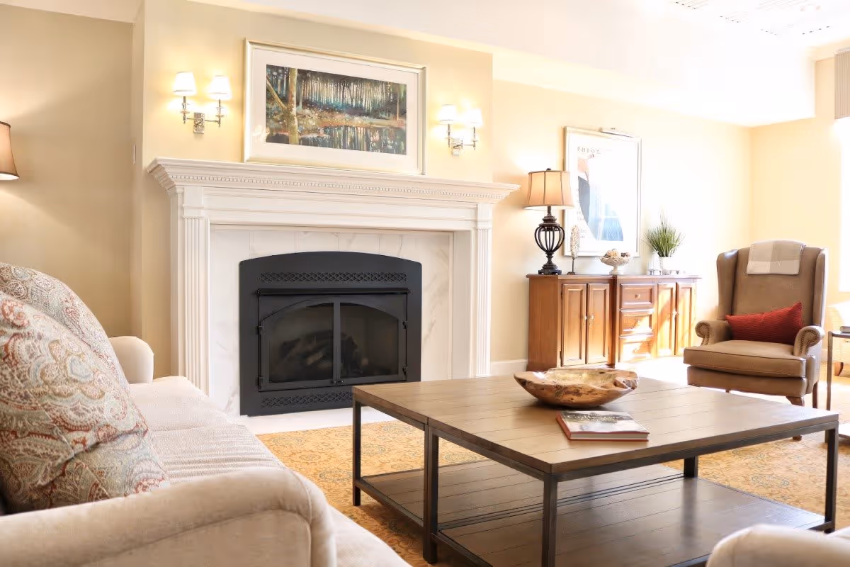 A cozy living room with a beige sofa adorned with patterned pillows, a wooden coffee table with a decorative bowl and a book, a fireplace with a white mantel, wall sconces, a wooden sideboard with a lamp and decor, and a comfortable armchair with a red cushion and a throw blanket.