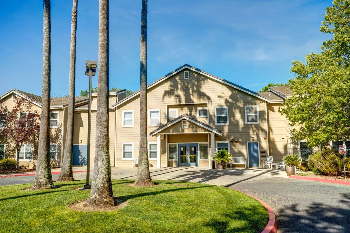 Exterior view of a two-story beige senior living facility building with a peaked roof, multiple windows, and a covered entrance. The foreground features a circular driveway with green grass and tall palm trees, along with benches and potted plants near the entrance under a clear blue sky.