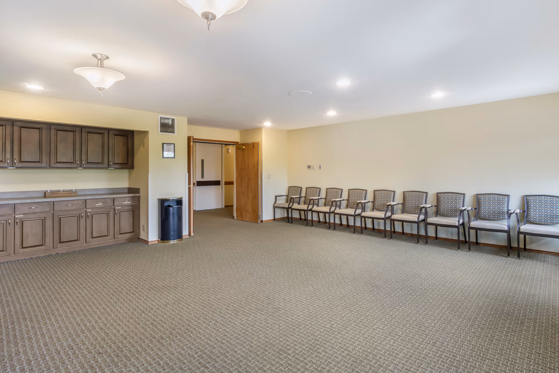Large empty multipurpose room with a row of chairs along the right wall, built-in cabinets on the left, and an open carpeted floor.