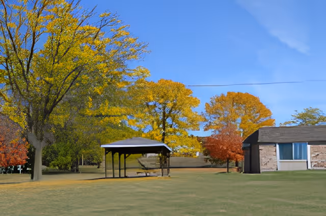 Open grassy area with a small covered pavilion, colorful autumn trees, and part of a single-story building.
