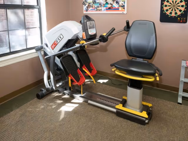 A SciFit exercise machine with a seat and foot pedals in a room with brown walls and carpeted floor. There is a window on the left side letting in natural light, a dartboard on the wall to the right, and a bulletin board with photos above the machine.