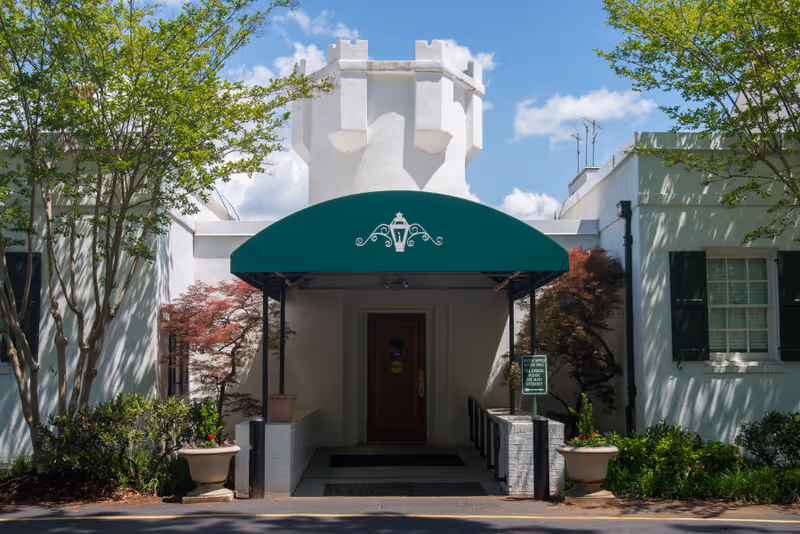 Entrance of a white stucco building with a green awning over a wooden door, flanked by planters and trees.