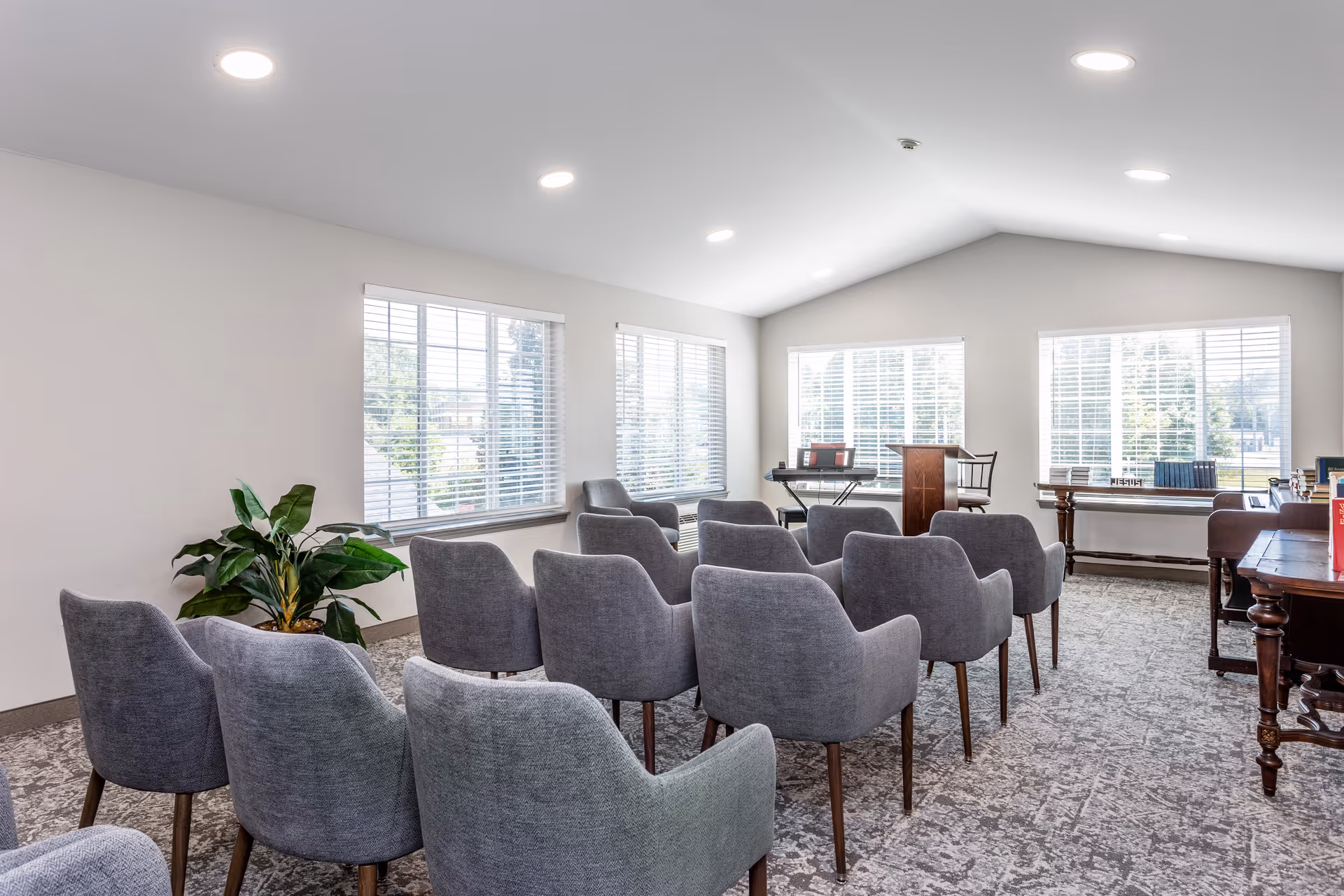A bright room with multiple gray upholstered chairs arranged in rows facing a wooden podium and a keyboard. Large windows with white blinds let in natural light, and there are tables with books and a plant in the corner.