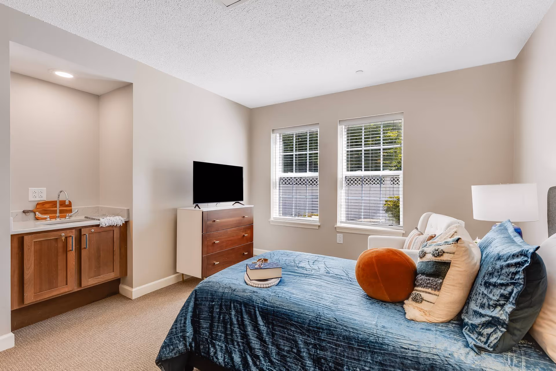 Well-lit bedroom with a blue-covered bed, decorative pillows, a dresser with a TV, a small sink cabinet, and two windows.
