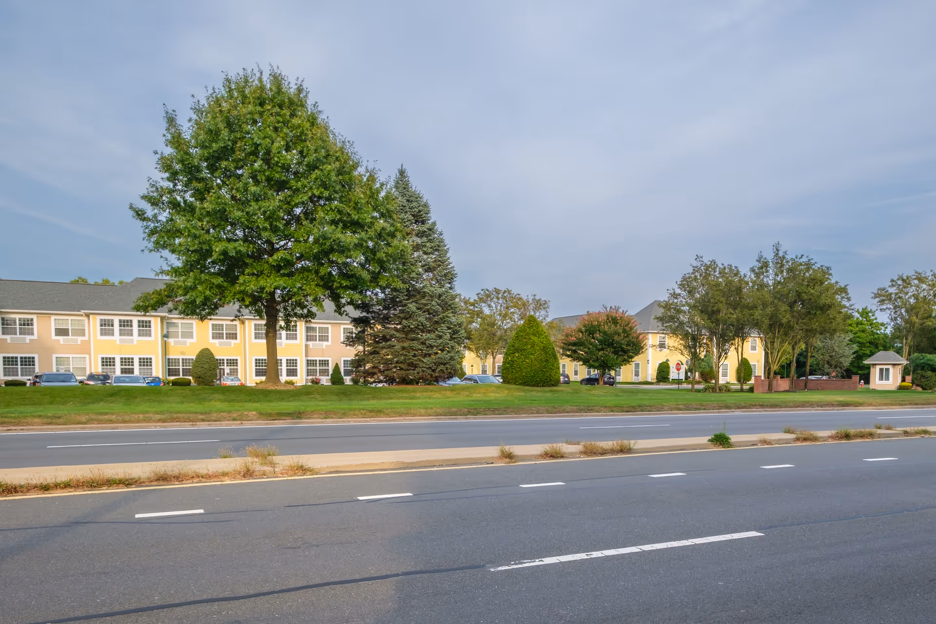 Exterior view of Brandywine Huntington Terrace by Monarch, showing a two-story yellow building with multiple windows, surrounded by green trees and bushes, with a road in the foreground.
