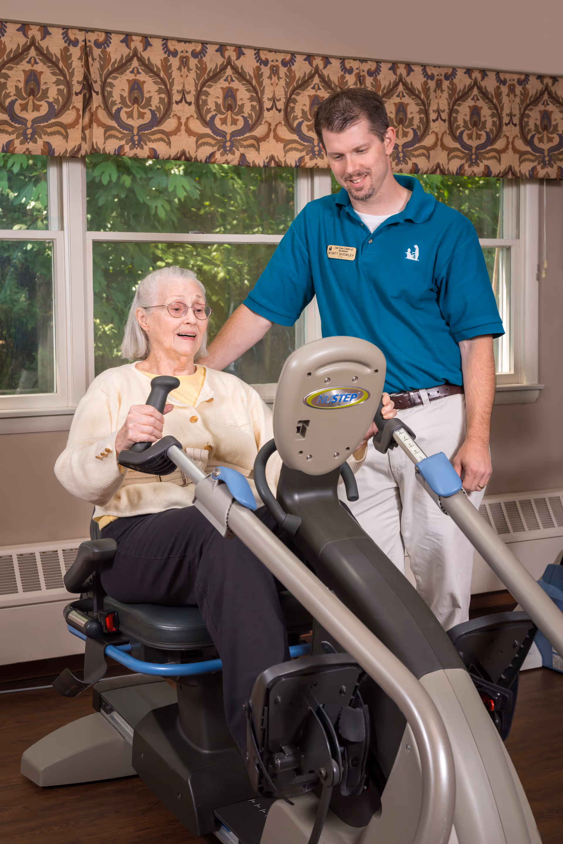 An elderly woman is seated on a NuStep exercise machine, holding the handles, while a male staff member in a teal polo shirt stands beside her, offering support and guidance. They are indoors in a room with large windows and patterned valances, with greenery visible outside.
