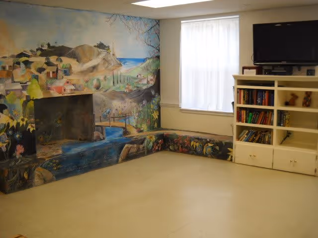 A cozy room with a painted fireplace mural depicting a scenic landscape with hills, water, and flowers. There is a window with white curtains letting in natural light. A white bookshelf filled with books and decorative items is positioned under a mounted flat-screen TV.