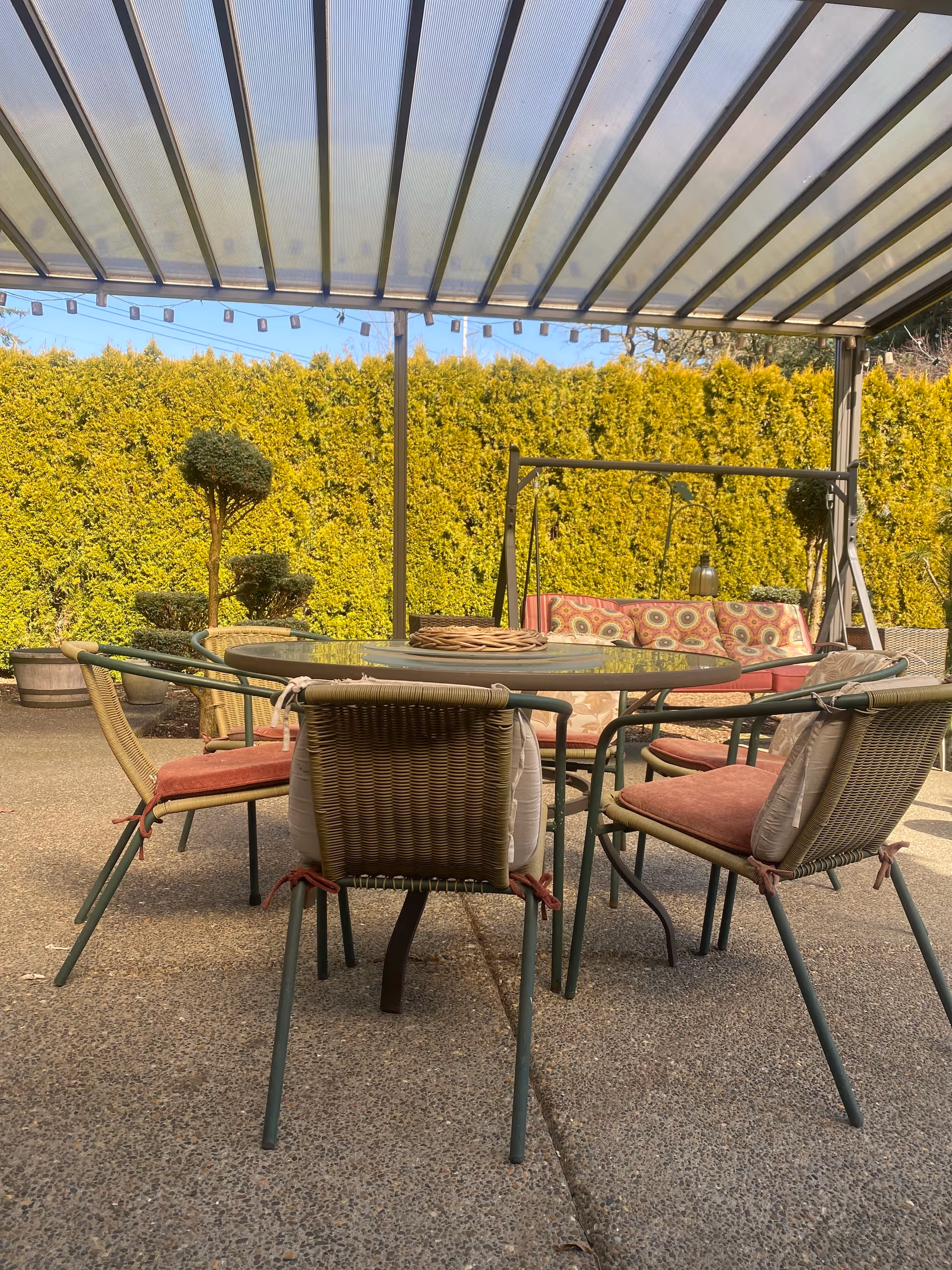 Outdoor patio area with a glass-top round table surrounded by six wicker chairs with red cushions. Behind the table is a cushioned swing bench with patterned pillows. The area is covered by a translucent roof and bordered by tall green hedges.