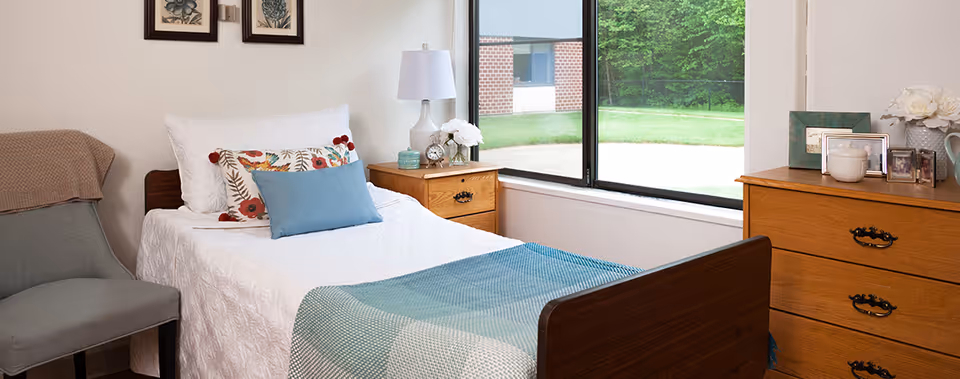 A neatly made single bed with white bedding, a blue throw blanket, and decorative pillows in a bright room. Next to the bed is a wooden nightstand with a white lamp, a clock, and a small vase of flowers. A wooden dresser with framed photos and a flower arrangement is on the right side. A gray chair with a beige throw blanket is positioned on the left. A large window shows a view of green grass and trees outside.