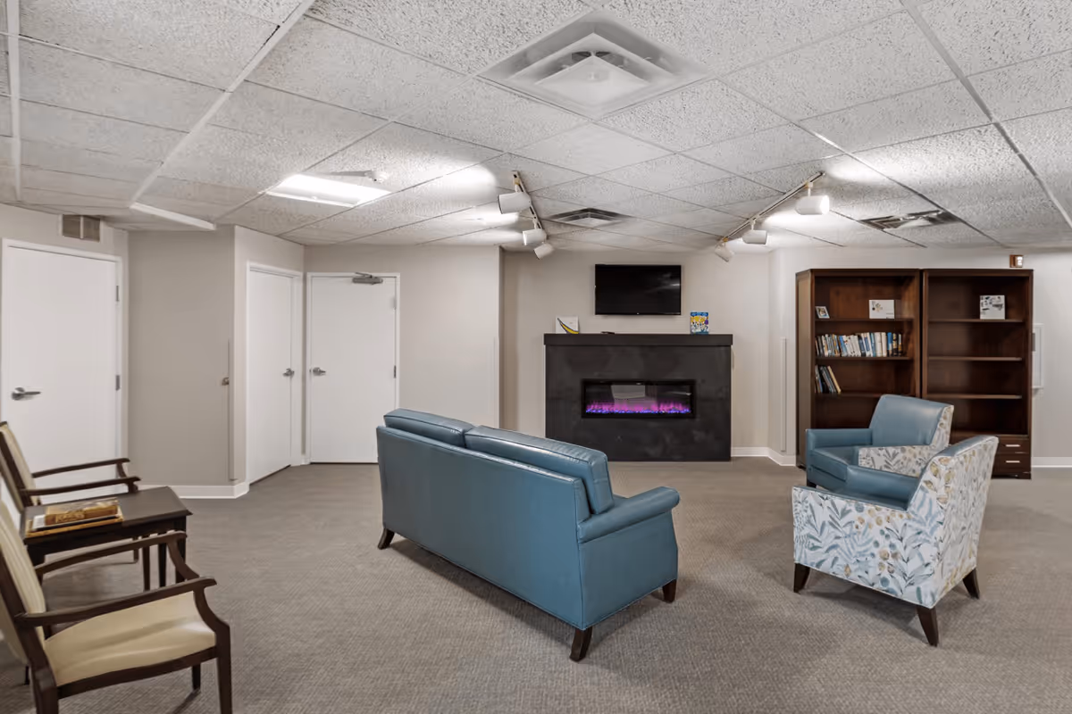 A cozy senior living common area with a blue leather sofa, two armchairs—one blue and one floral patterned—facing a black electric fireplace with purple flames. Above the fireplace is a mounted flat-screen TV. To the right, there are two wooden bookshelves with books and decorative items. On the left side, there are two wooden chairs with beige cushions and a small table between them. The room has a carpeted floor, beige walls, and a white drop ceiling with fluorescent and track lighting.