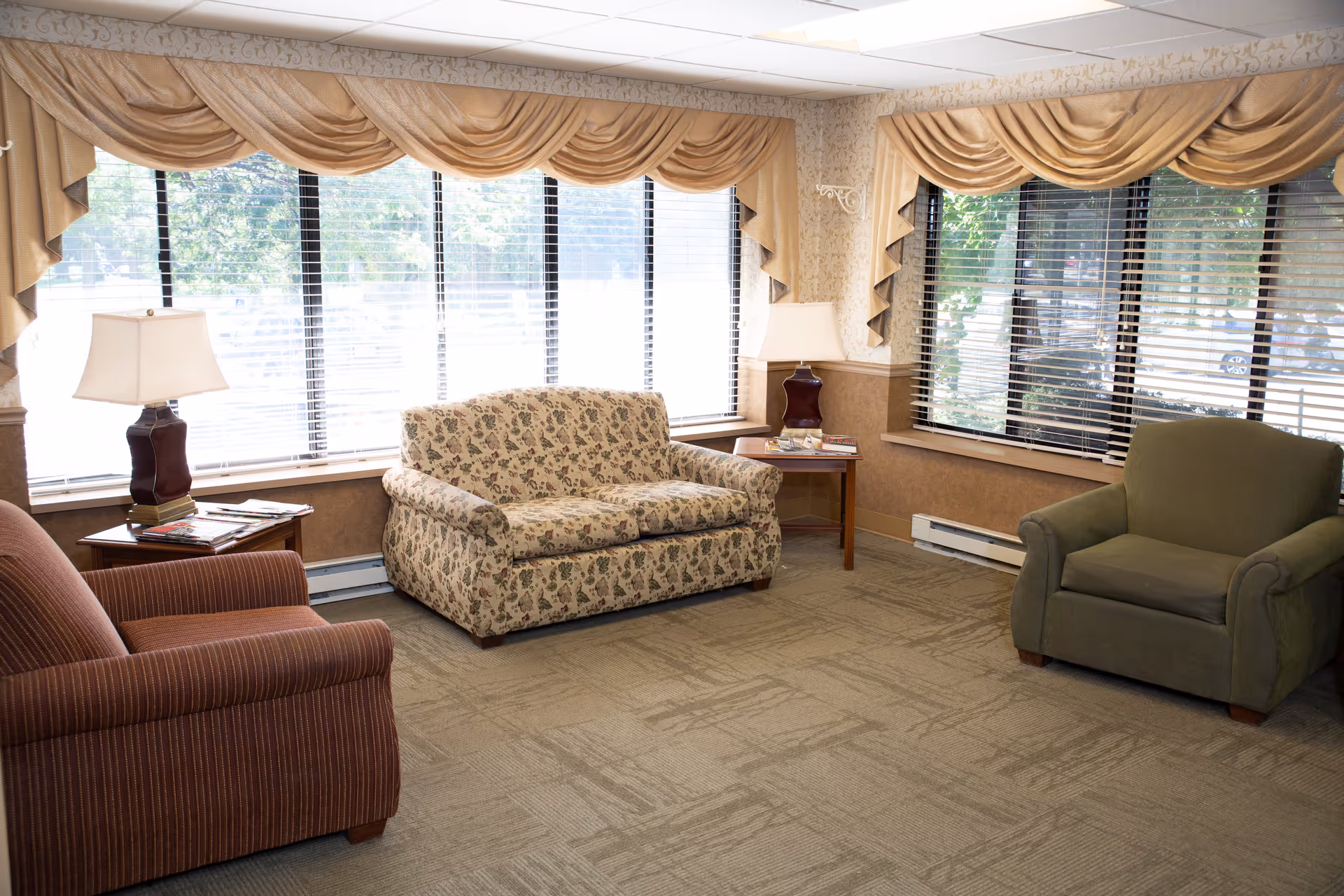 A bright sitting room with upholstered chairs and a floral loveseat arranged near large windows with blinds and draped valances.