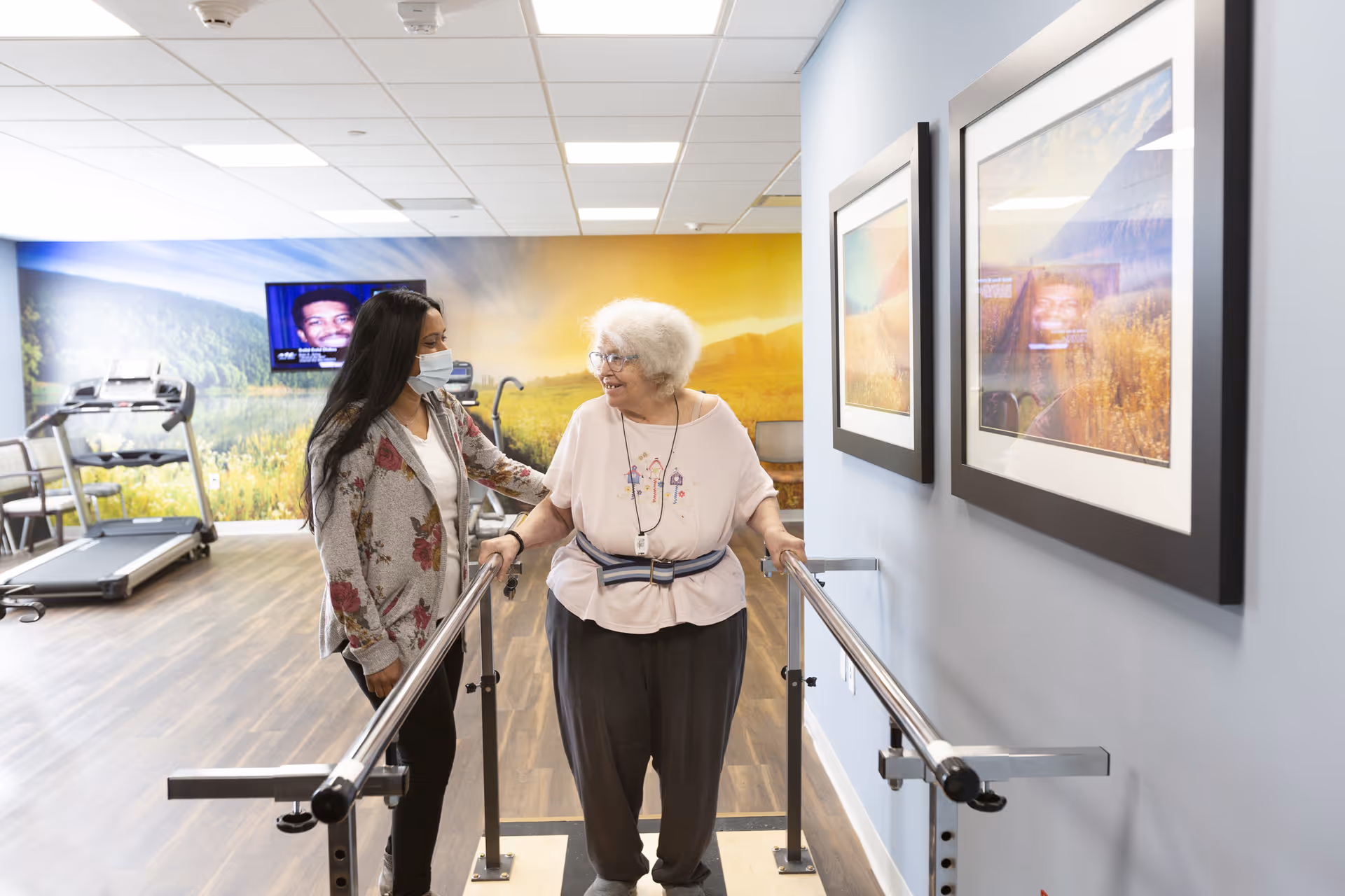 An elderly woman using parallel bars for walking exercise is assisted by a caregiver wearing a face mask in a bright rehabilitation or fitness room with treadmills and a large scenic mural on the wall.