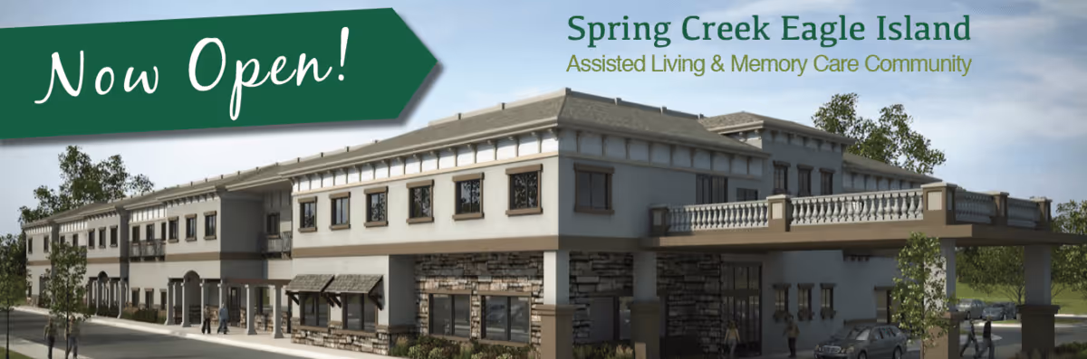 Exterior view of a two-story assisted living and memory care community building with stone and stucco facade, several windows, and a covered entrance. There are a few people walking near the building and cars parked nearby. A green banner in the top left corner reads 'Now Open!'.