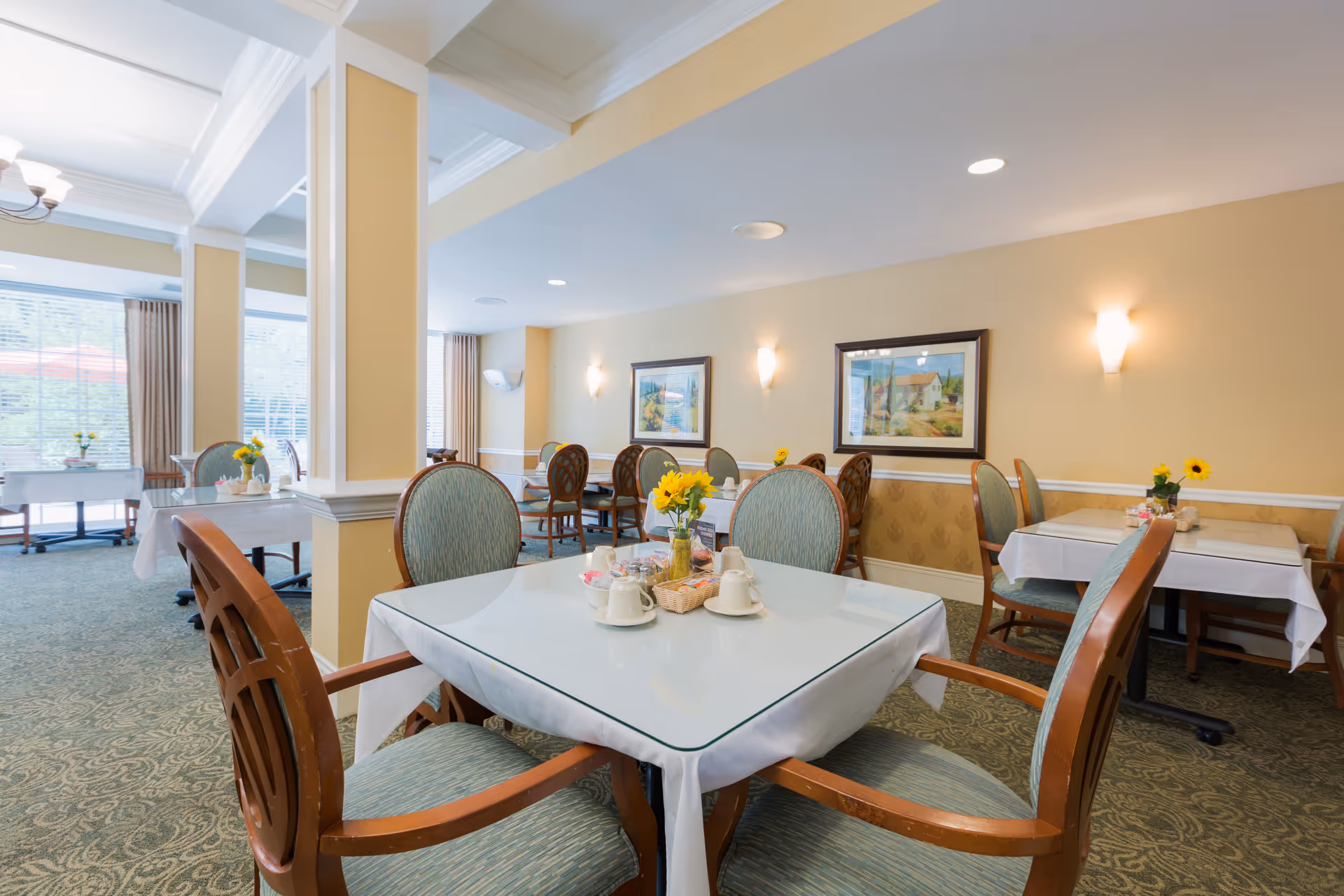 Bright dining room with multiple tables and chairs set with tableware and small flower centerpieces.