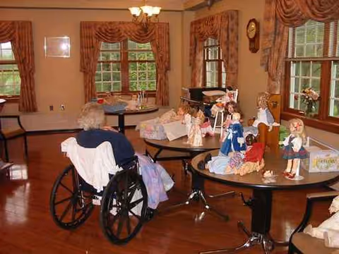 Elderly resident in a wheelchair sits at a round table in a common room with dolls.