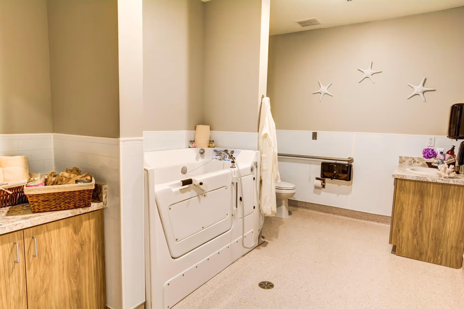 A spacious bathroom with a walk-in bathtub, a toilet with a grab bar, a sink with a countertop, and wooden cabinets. The walls are decorated with three white starfish, and there is a basket with towels and toiletries on the counter.