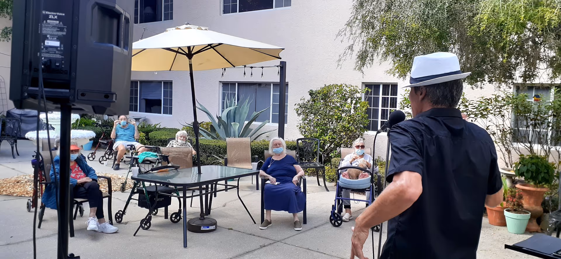 An outdoor patio area at a senior living facility where elderly residents are seated in chairs spaced apart, some using walkers. A man wearing a white hat and black shirt is speaking or performing into a microphone facing the residents. There is a large speaker on a stand to the left and a table with an umbrella in the center. The background shows a building with windows and some potted plants.