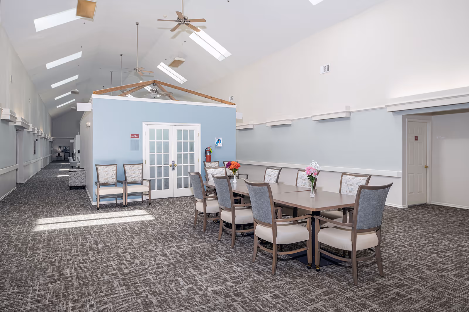 A spacious senior living facility common area with a long table surrounded by chairs. The room has high ceilings with skylights and ceiling fans. There are two small flower vases on the table and additional chairs against a light blue wall with double glass doors. The carpet has a patterned design and the walls are painted in light colors.