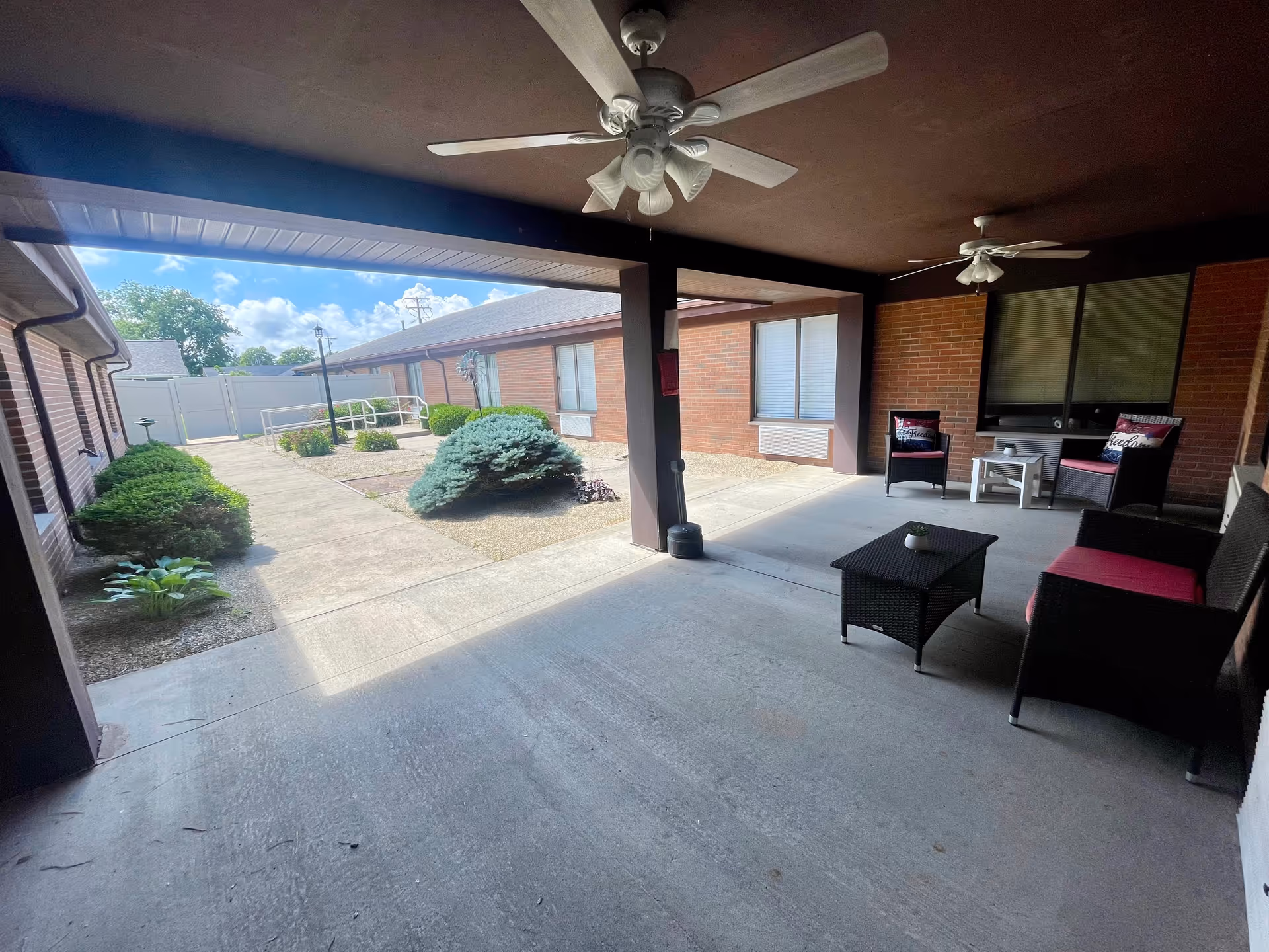 Covered outdoor patio area with ceiling fans, wicker seating with red cushions, a coffee table, and a small side table. The patio overlooks a courtyard with concrete walkways, bushes, and a small tree, surrounded by brick buildings under a blue sky with clouds.