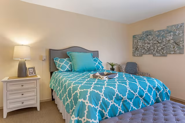 A bedroom with a bed featuring a turquoise and white patterned comforter and matching pillows. There is a white nightstand with a lamp and a framed photo on the left side of the bed. A decorative wall art piece with floral designs hangs on the beige wall above a patterned armchair. A tufted bench is positioned at the foot of the bed.